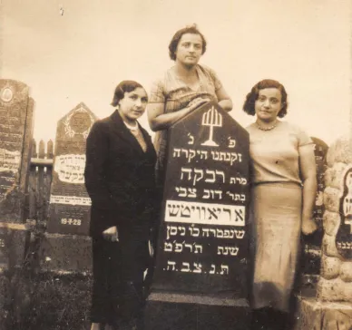 Photo of three women. They are the sisters of Harvey Sohnen’s grandmother. Rezia was a teacher, Nechama a housewife, and Lea owned a grocery. They were all murdered during the Holocaust. They stand near their mother’s grave in the Jewish graveyard of Ashmyany.  Rivka Aryewitz, daughter of Dov Tsvi Oriavitsh, died in May 1929. She was likely the great-aunt of Harvey’s grandmother Hannah.   The photo was taken between May 1929 and the Second World War. On the back of the photo, a text written in Yiddish says: