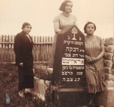 Photo of three women. They are the sisters of Harvey Sohnen’s grandmother. Rezia was a teacher, Nechama a housewife, and Lea owned a grocery. They were all murdered during the Holocaust. They stand near their mother’s grave in the Jewish graveyard of Ashmyany. Rivka Aryewitz, daughter of Dov Tsvi Oriavitsh, died in May 1929. She was likely the great-aunt of Harvey’s grandmother Hannah.  The photo was taken between May 1929 and the Second World War. On the back of the photo, a text written in Yiddish says:  