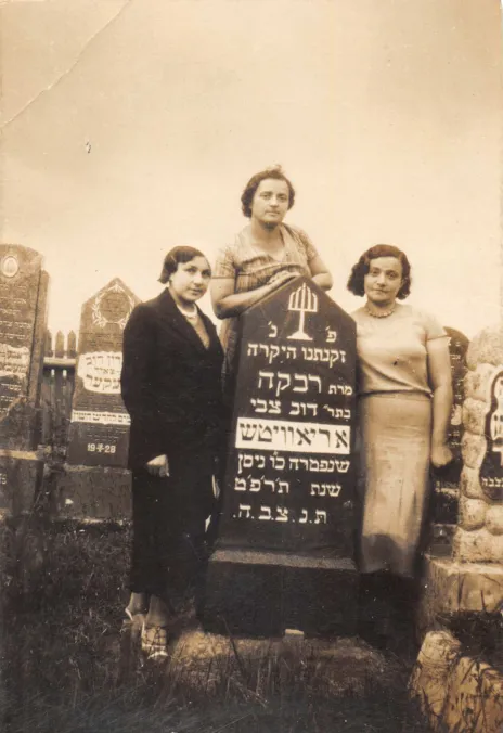 Photo of three women. They are the sisters of Harvey Sohnen’s grandmother. Rezia was a teacher, Nechama a housewife, and Lea owned a grocery. They were all murdered during the Holocaust. They stand near their mother’s grave in the Jewish graveyard of Ashmyany.  Rivka Aryewitz, daughter of Dov Tsvi Oriavitsh, died in May 1929. She was likely the great-aunt of Harvey’s grandmother Hannah.   The photo was taken between May 1929 and the Second World War. On the back of the photo, a text written in Yiddish says: