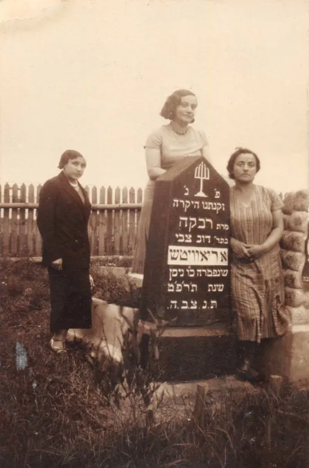 Photo of three women. They are the sisters of Harvey Sohnen’s grandmother. Rezia was a teacher, Nechama a housewife, and Lea owned a grocery. They were all murdered during the Holocaust. They stand near their mother’s grave in the Jewish graveyard of Ashmyany. Rivka Aryewitz, daughter of Dov Tsvi Oriavitsh, died in May 1929. She was likely the great-aunt of Harvey’s grandmother Hannah.  The photo was taken between May 1929 and the Second World War. On the back of the photo, a text written in Yiddish says:  